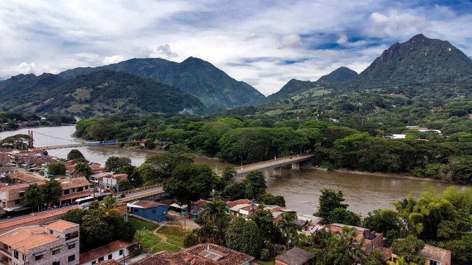 A river with a bridge, a town with red-roofed buildings, and green mountains in the background.