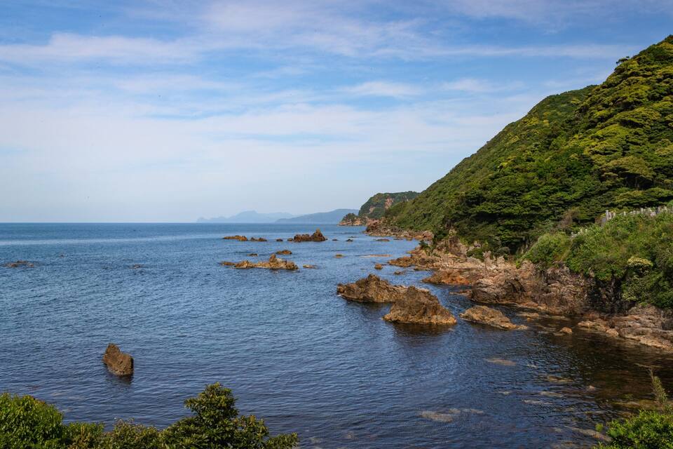A coastal landscape with rocky outcrops in the water, a green hillside, and a clear sky.