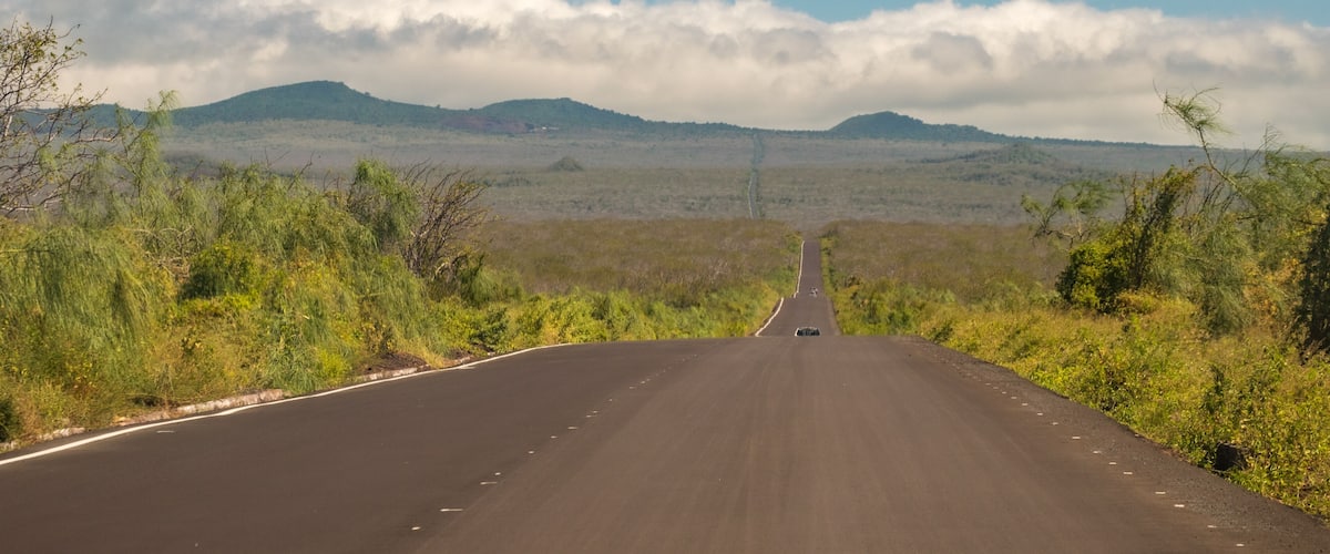 A long, straight road with a vehicle in the distance, surrounded by sparse vegetation and hills under a partly cloudy sky.