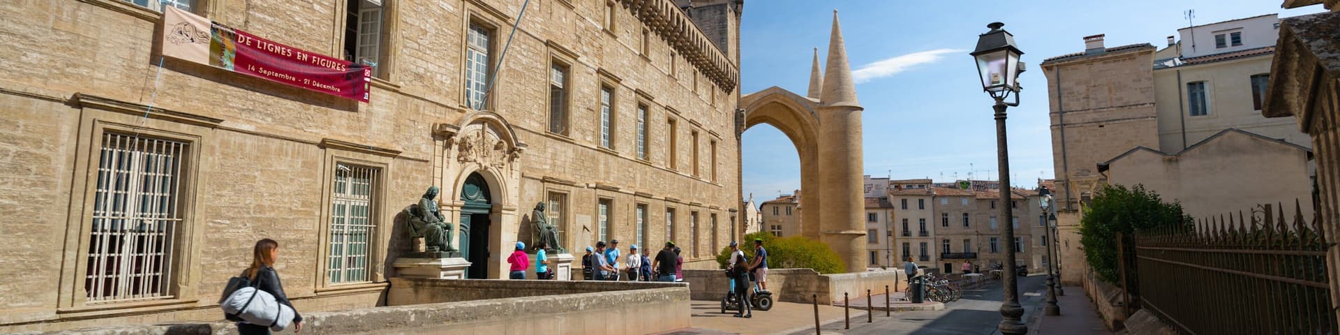 Un edificio histórico con una estatua, una torre y una calle con gente caminando.