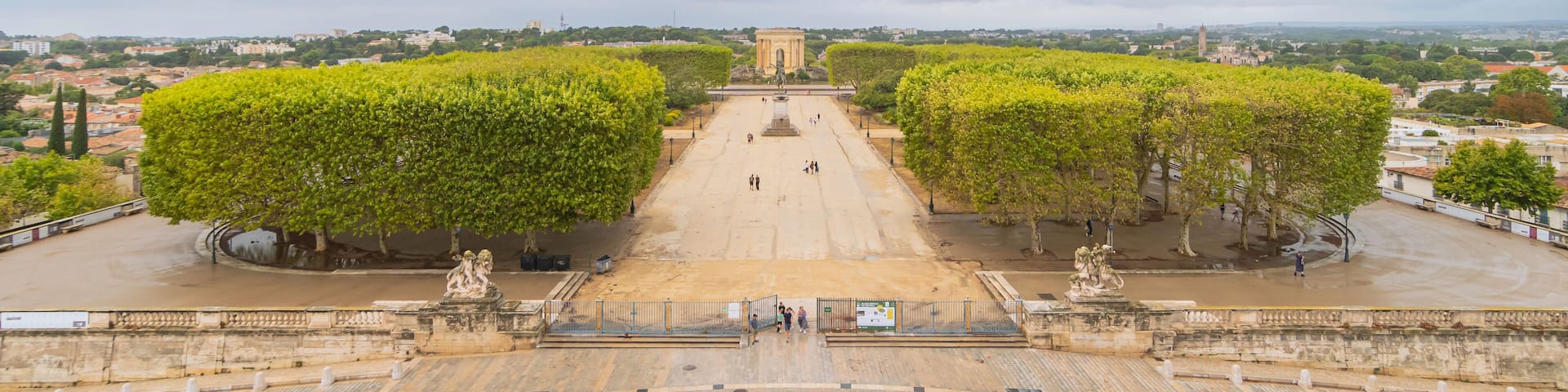 A wide plaza with a central monument, surrounded by trees and a distant cityscape under cloudy skies.