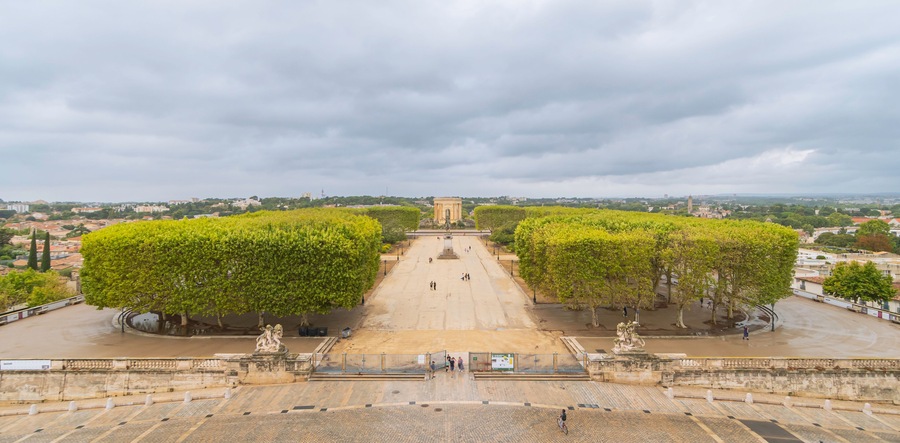 Une vaste place, ornée d’un monument central, entourée d’arbres et offrant une vue lointaine sur la ville, le tout sous un ciel nuageux.