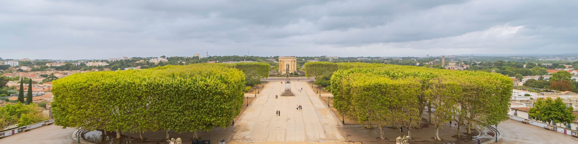 Una amplia plaza con un monumento central, rodeada de árboles y un horizonte urbano lejano bajo cielos nublados.