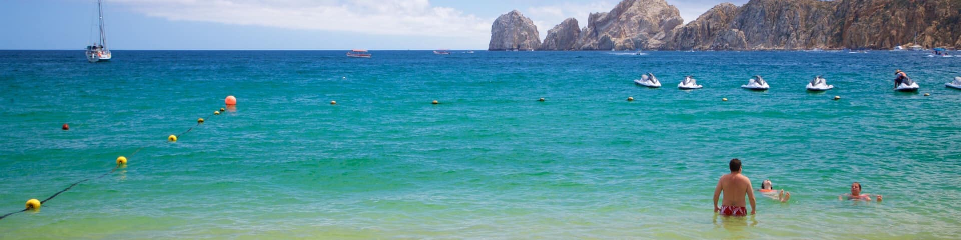 A beach with clear turquoise water, a sailboat, and rocky cliffs in the background.
