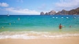 A beach with clear turquoise water, a sailboat, and rocky cliffs in the background.