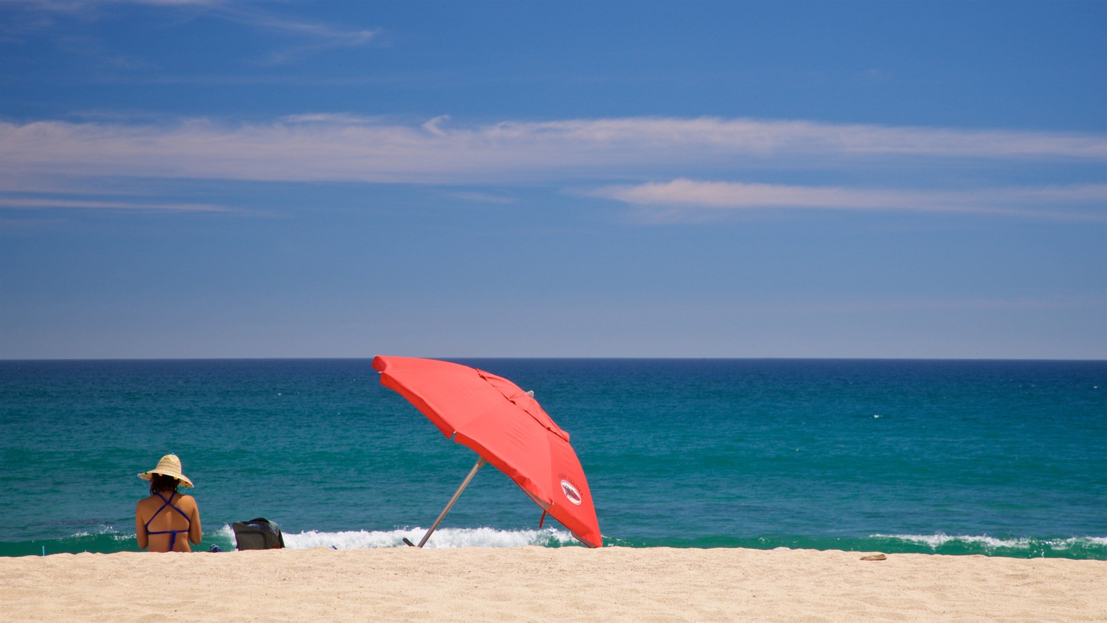 Ein Strand mit einem roten Sonnenschirm, einer Frau mit Strohhut und dem Meer.