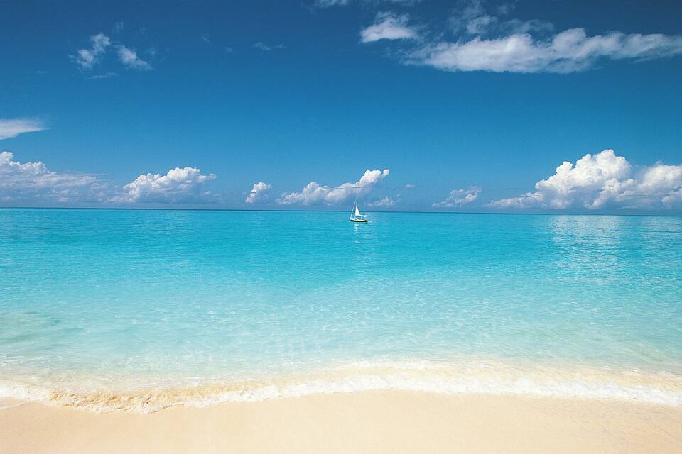 Une mer turquoise aux eaux limpides, avec un voilier blanc à l’horizon et une plage de sable au premier plan.