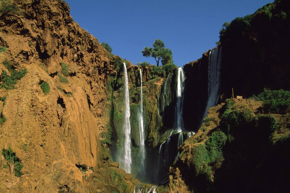 A multi-tiered waterfall surrounded by rocky terrain and sparse vegetation.