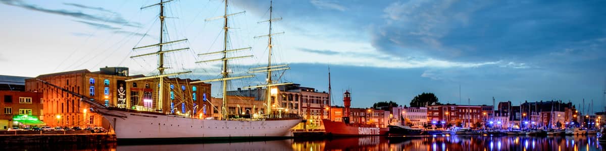 A historic sailing ship docked at a waterfront with buildings and a lighthouse in the background.