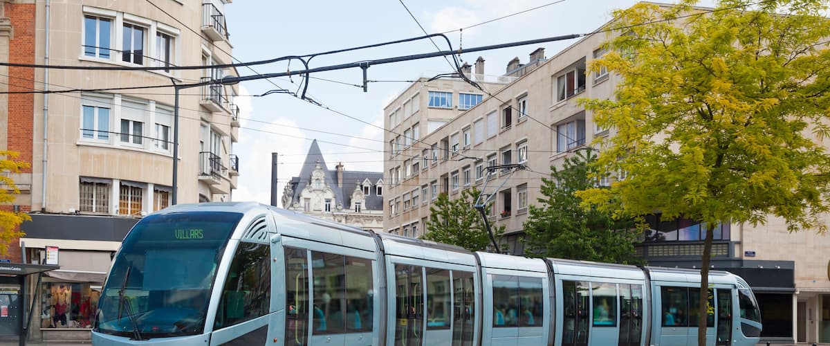 A modern light blue tram on tracks in an urban setting with multi-story buildings and a tree with yellow leaves.
