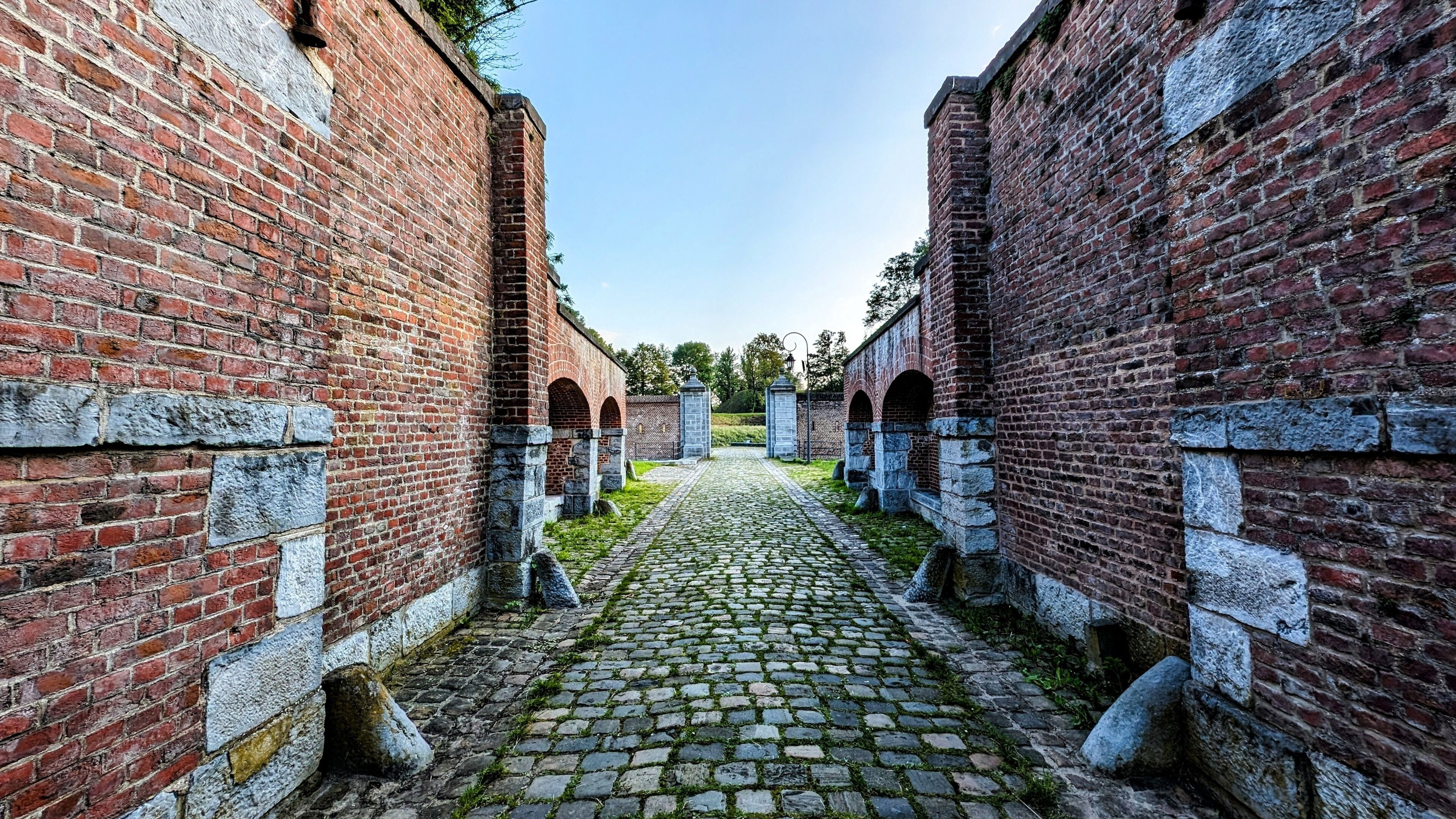 A cobblestone pathway flanked by red brick walls with stone pillars and arched openings.