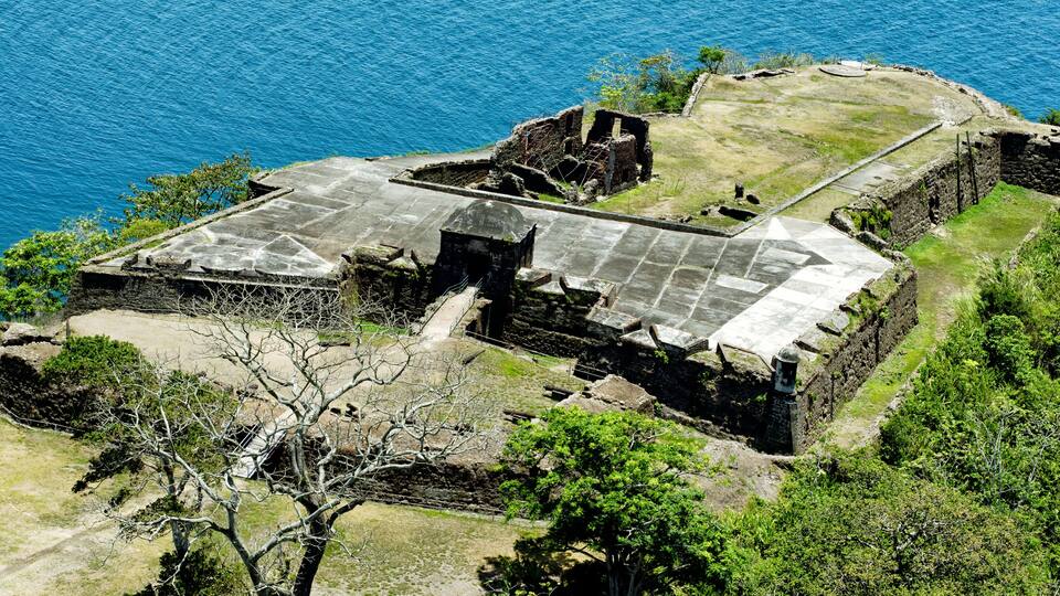 An ancient stone structure with a flat roof, surrounded by lush greenery and overlooking a body of water.