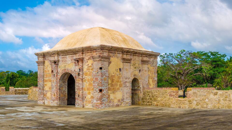 A historic stone structure with a dome and arched entrance, surrounded by greenery and palm trees.