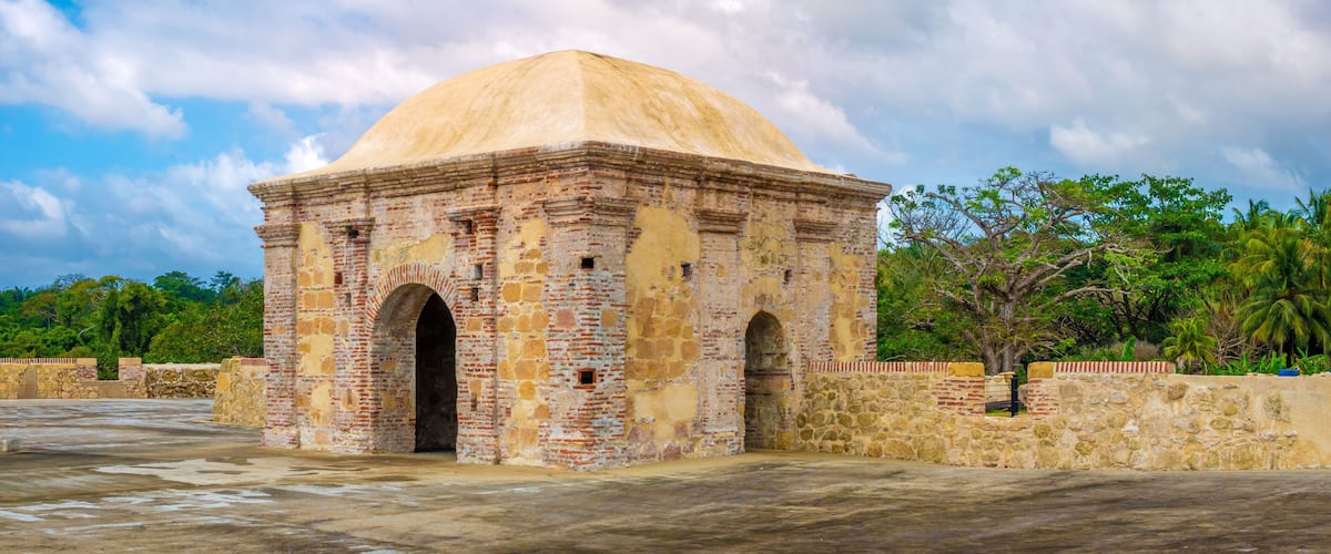 A historic stone structure with a dome and arched entrance, surrounded by greenery and palm trees.