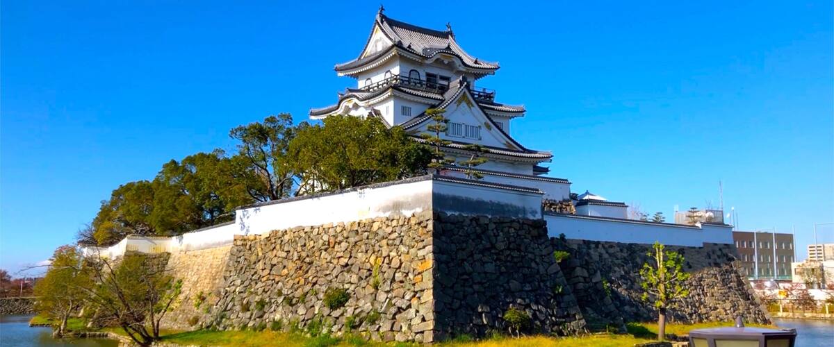 A traditional Japanese castle with a stone base and multiple tiers, surrounded by a moat and greenery.