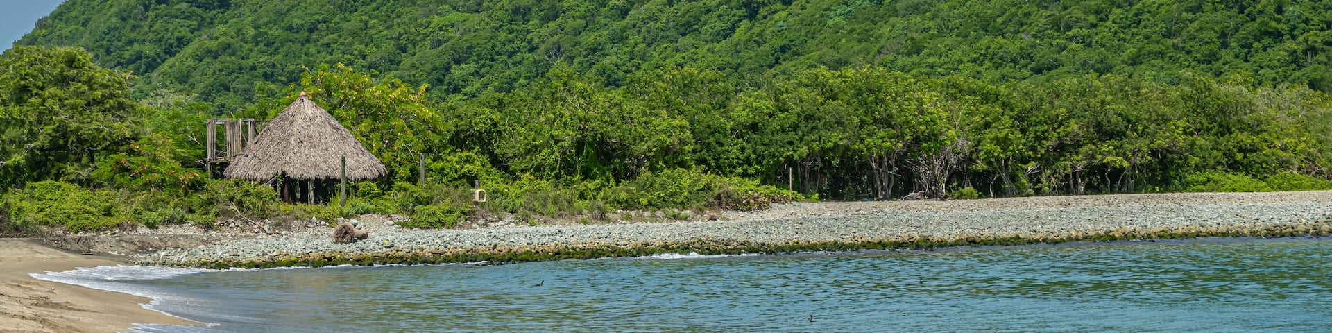 Zihuatanejo, Mexico - July 18, 2023: Green forested hill on Punta Potosi at south end of Playa Larga. Traditional hut just off beach half hidden in jungle. Ocean water and sand. Blue sky