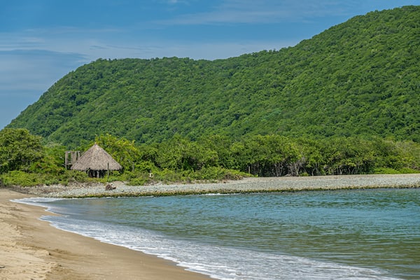 Zihuatanejo, Mexico - July 18, 2023: Green forested hill on Punta Potosi at south end of Playa Larga. Traditional hut just off beach half hidden in jungle. Ocean water and sand. Blue sky