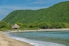Zihuatanejo, Mexico - July 18, 2023: Green forested hill on Punta Potosi at south end of Playa Larga. Traditional hut just off beach half hidden in jungle. Ocean water and sand. Blue sky