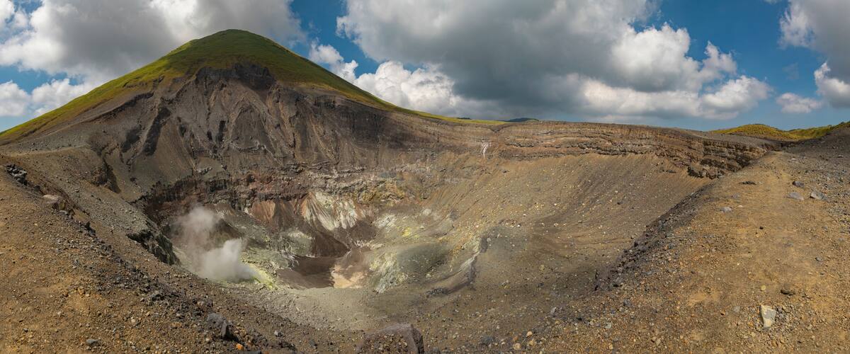 A volcanic crater with a green hill in the background and a cloudy sky.