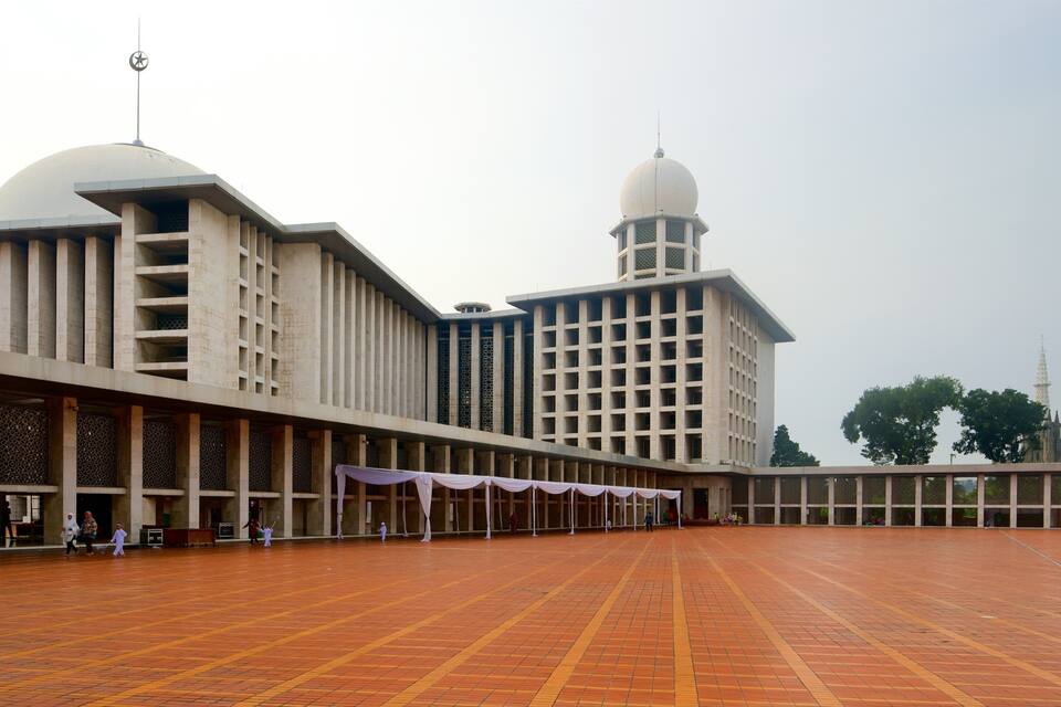 A large, modern building with a dome and a minaret, surrounded by a spacious courtyard with patterned flooring.