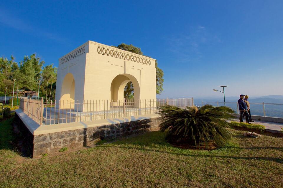 A white monument with arches and a fence, surrounded by grass and trees, with a clear sky and distant mountains.