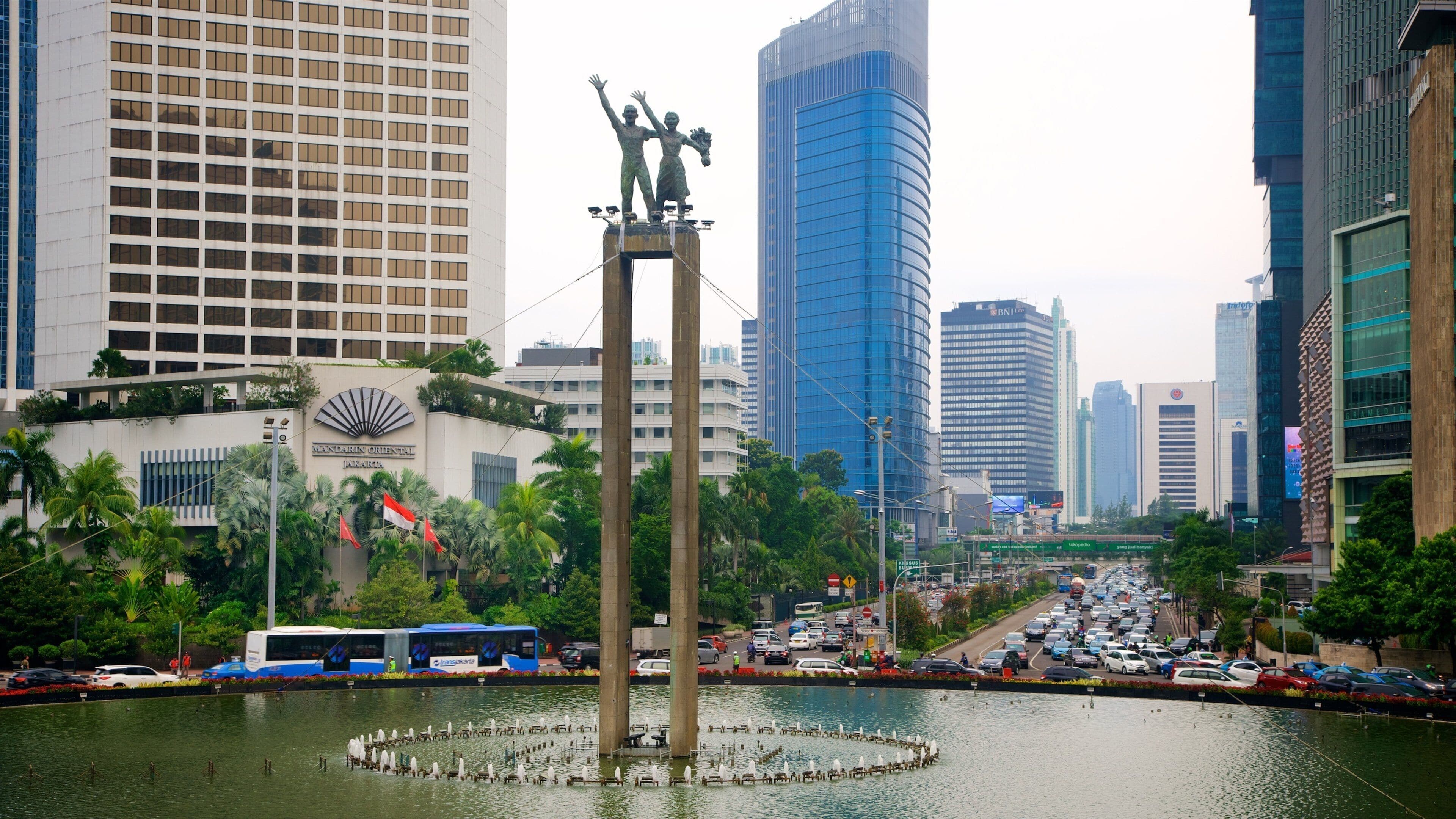 Eine Stadtansicht mit einer markanten Statue in der Mitte, umgeben von Wasser, hohen Gebäuden und einer belebten Straße mit Fahrzeugen.