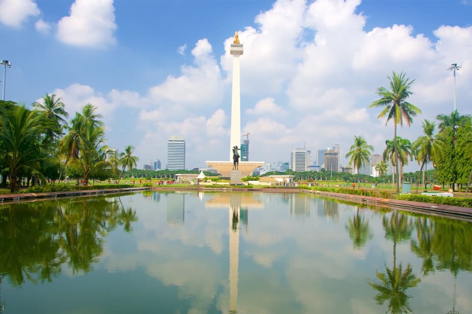 Um monumento alto com uma estátua no topo, rodeado por palmeiras e um corpo d'água que reflete o monumento e o céu.