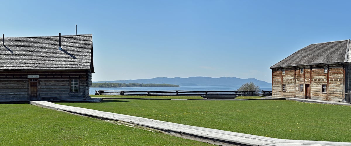 Two rustic log cabins with a wooden fence and a clear view of a lake and mountains in the background.