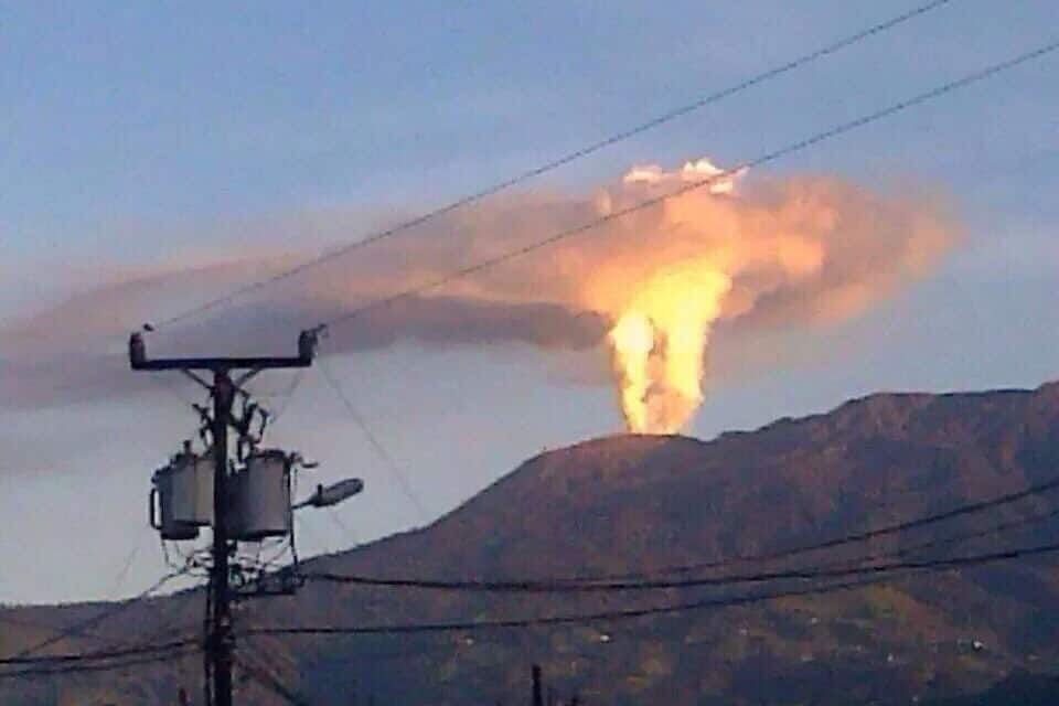 A volcano erupting with smoke and ash visible against a mountain backdrop.