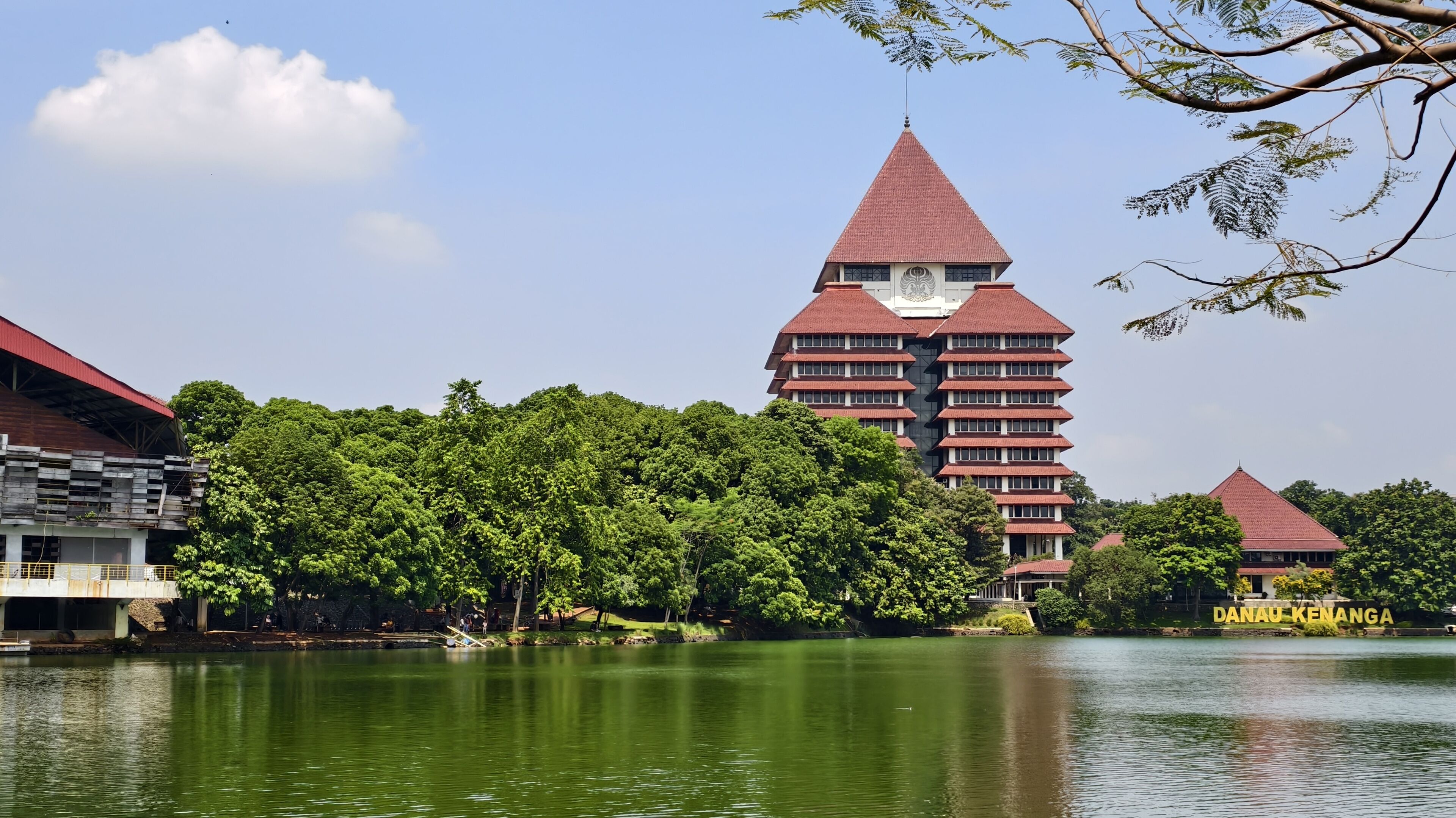 A traditional multi-tiered building with a red roof, surrounded by lush greenery and a calm lake.
