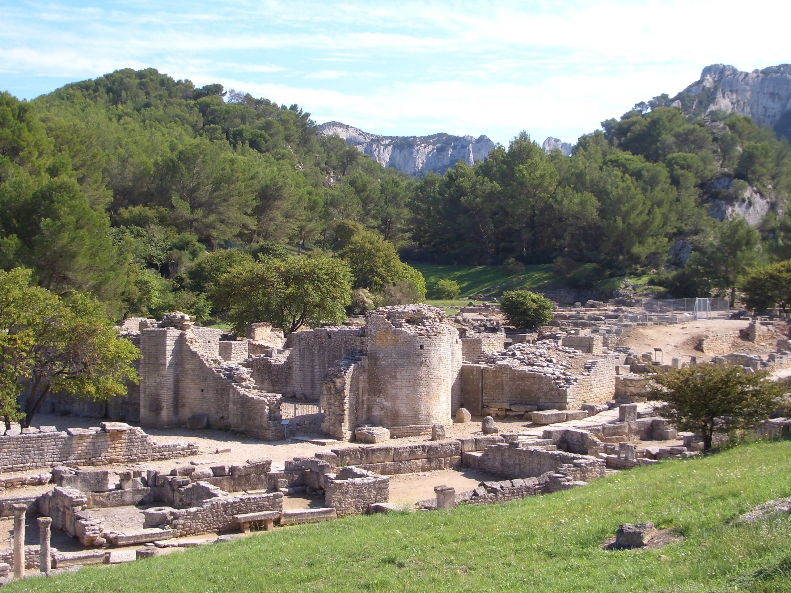 Ancient ruins with stone walls and columns, surrounded by lush green trees and mountains in the background.