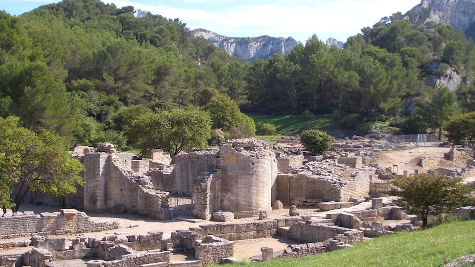 Ancient ruins with stone walls and columns, surrounded by lush green trees and mountains in the background.