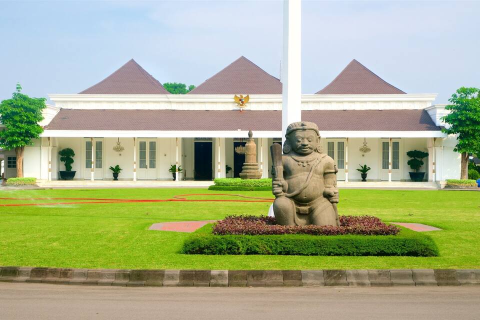 A building with a white facade and a red roof, a statue in the foreground, and a well-maintained lawn.