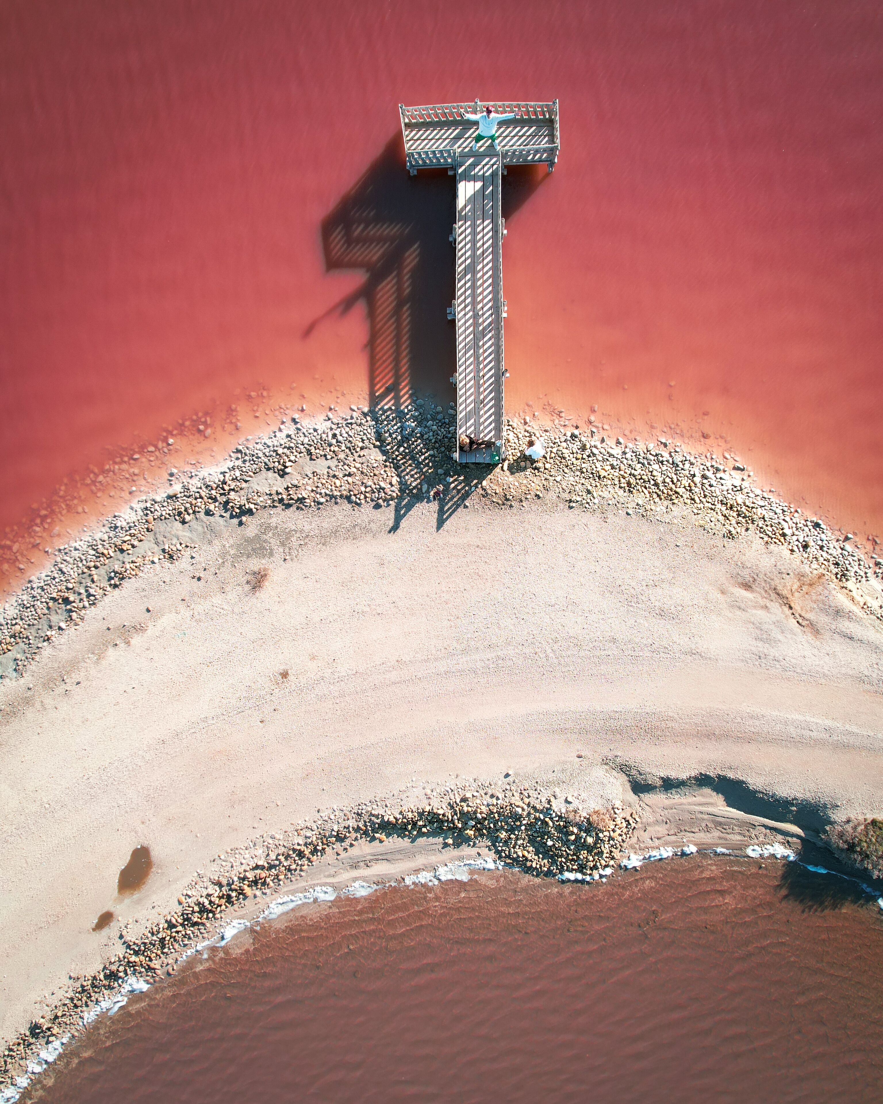 Aerial view of a wooden pier casting a long shadow on the vibrant pink waters of the salt flats, contrasting with the sandy beach, Arles, Provence-Alpes-Cote d'Azur, France.