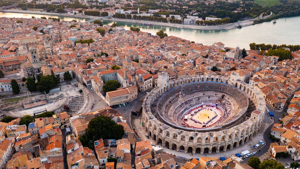 An aerial view of a historic city with a prominent ancient amphitheater, a river, and surrounding urban architecture.