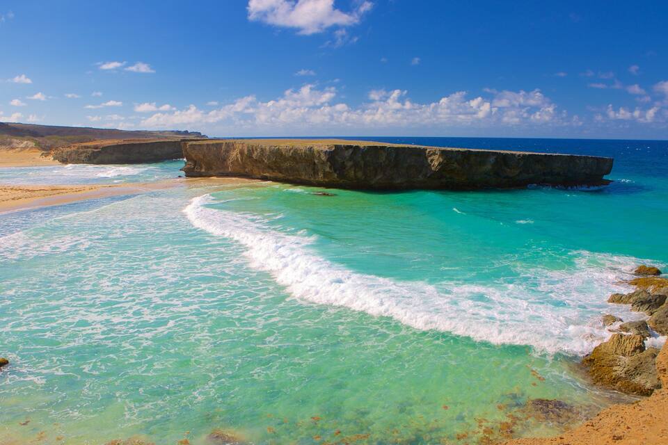 A coastal landscape with clear turquoise waters, a sandy beach, and a large rock formation.