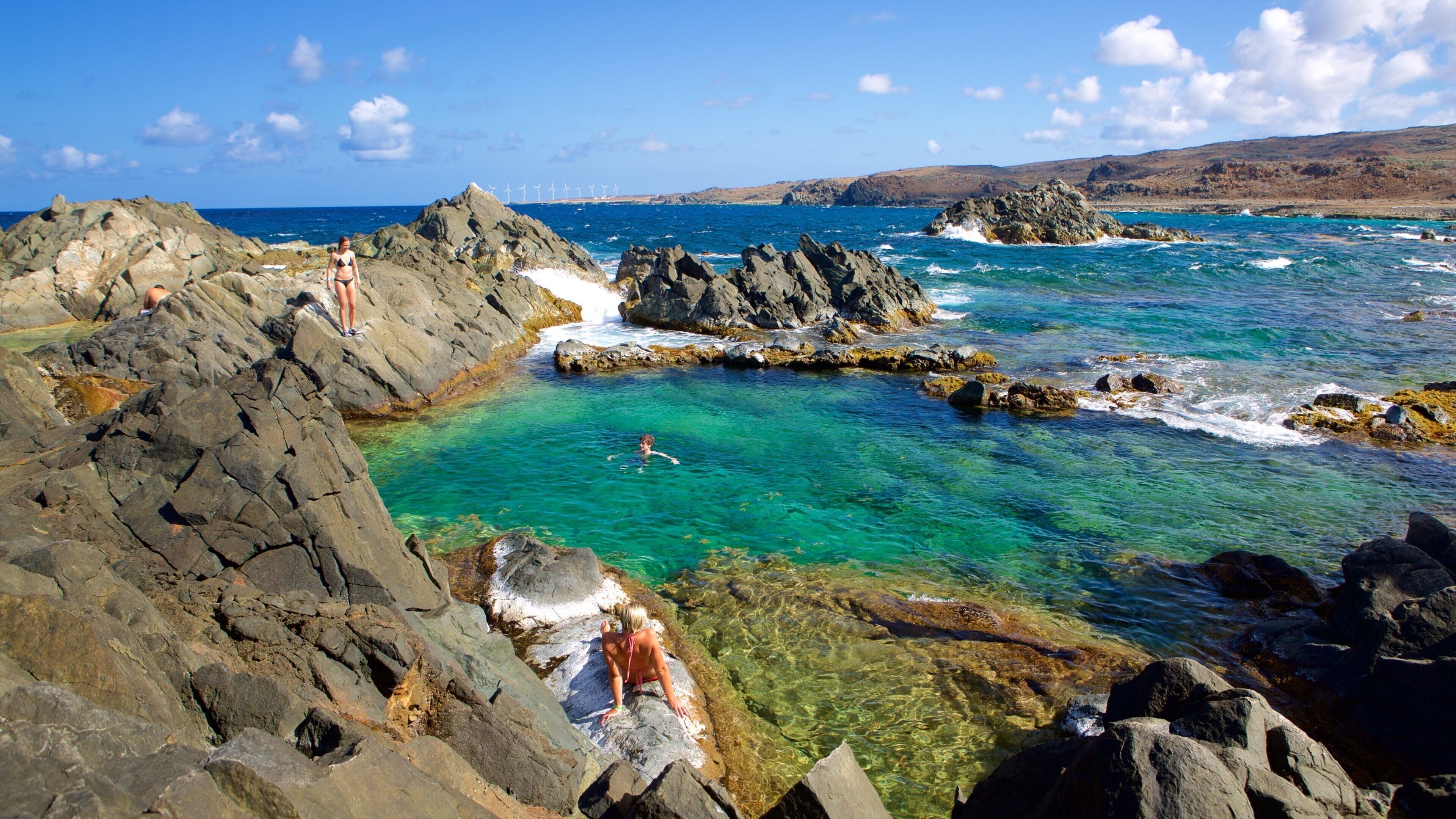 A rocky coastal landscape with clear turquoise waters, a person swimming, and a distant view of wind turbines.