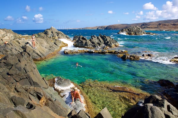 A rocky coastal landscape with clear turquoise waters, a person swimming, and a distant view of wind turbines.