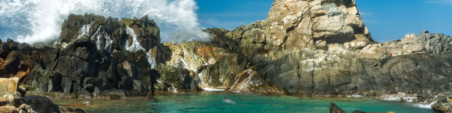 A rocky coastline with clear turquoise waters, a prominent rock formation, and a waterfall.