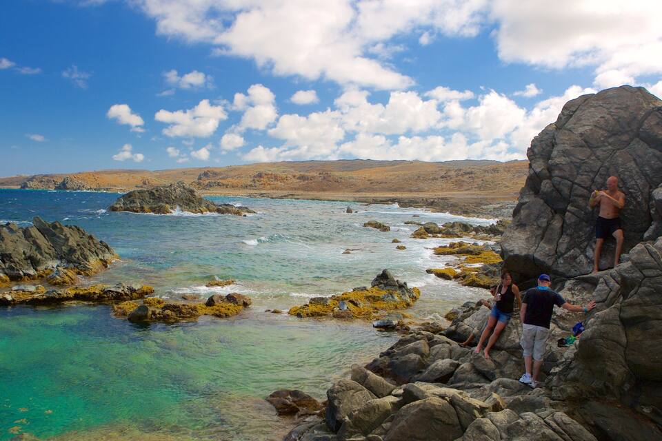 A rocky coastline with clear turquoise waters, a person climbing a rock, and another person standing nearby.