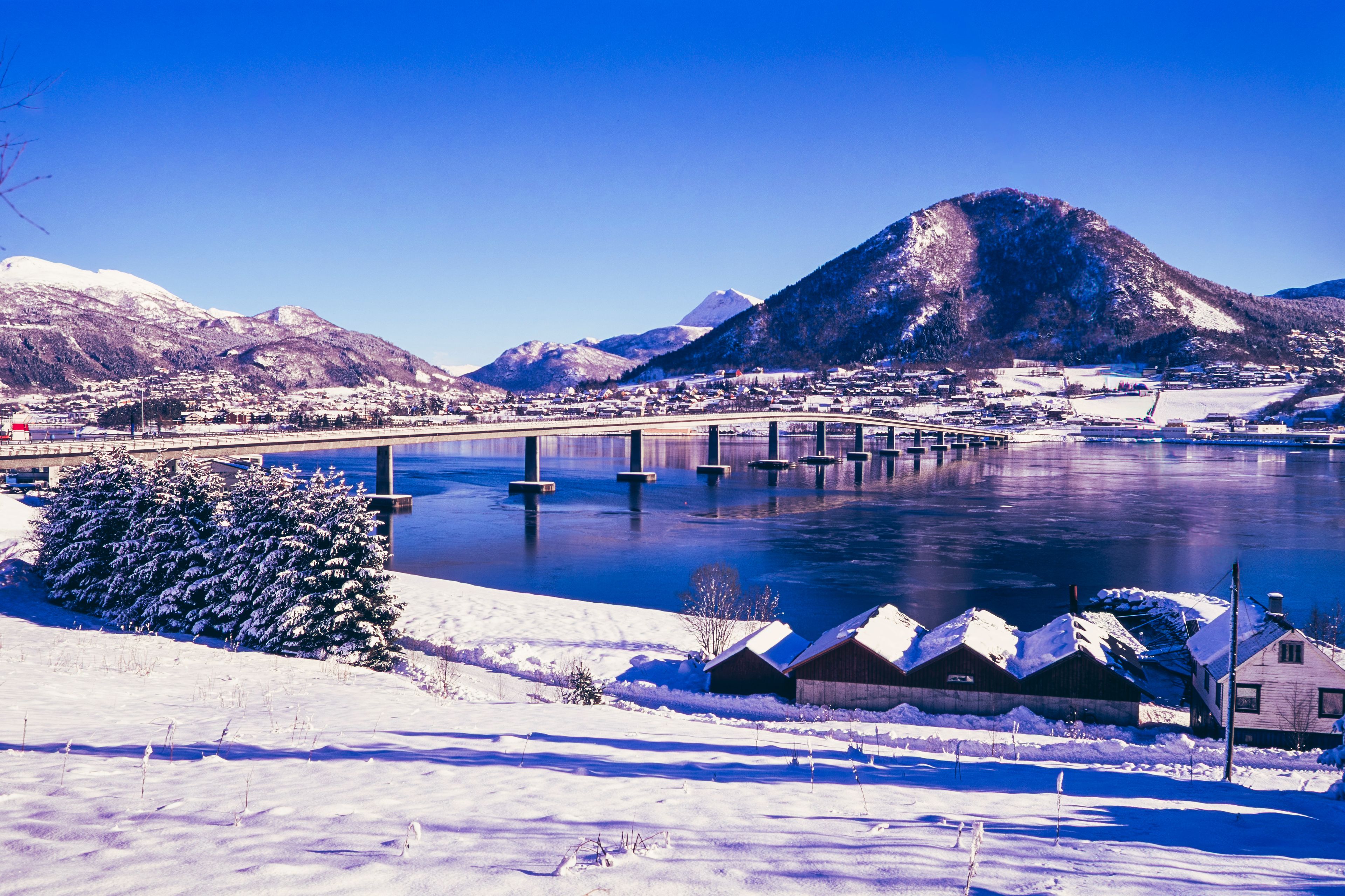 Paisagem coberta de neve com uma ponte sobre um rio, montanhas ao fundo e casas com neve nos telhados.