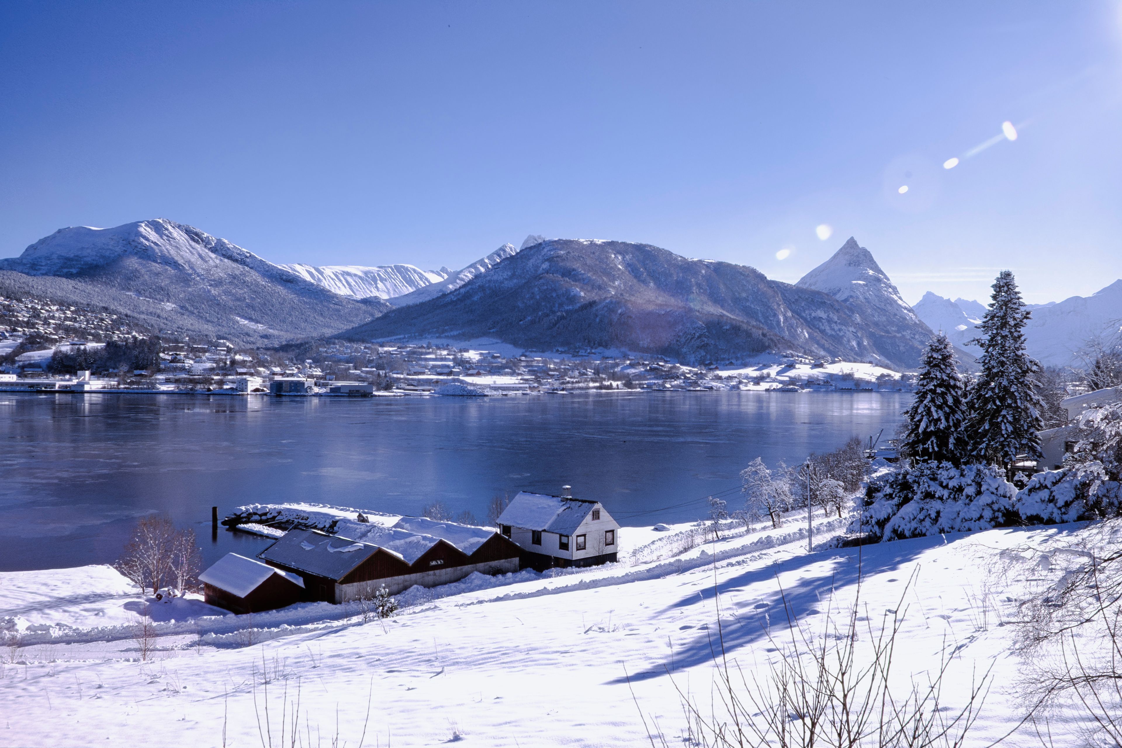 Paisagem coberta de neve com um lago, montanhas e uma fileira de casas.