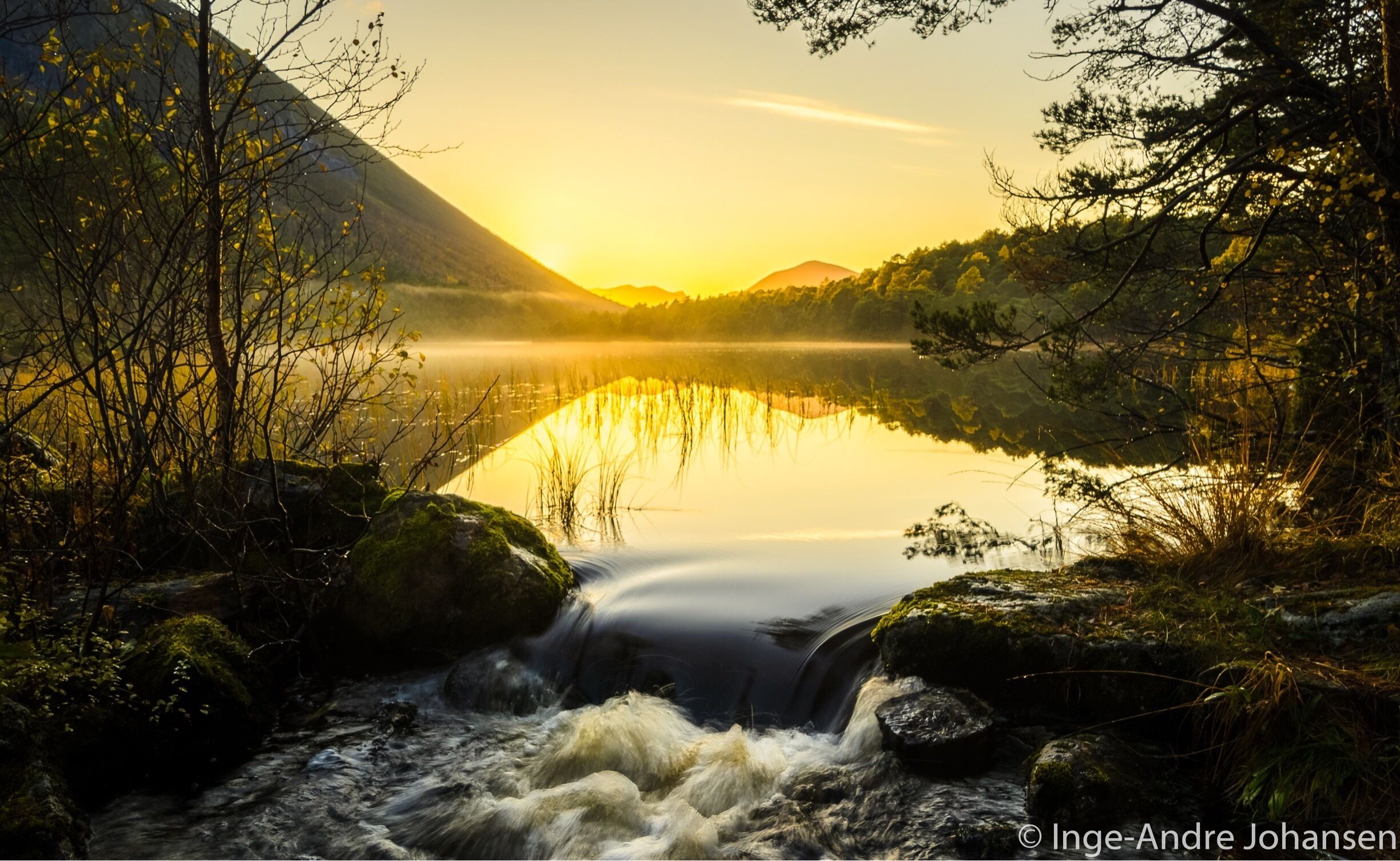 A calm lake reflecting the sunset, surrounded by trees and mountains.