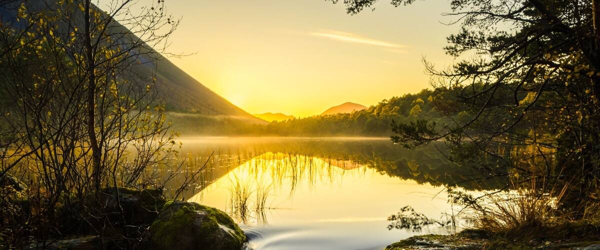 A calm lake reflecting the sunset, surrounded by trees and mountains.