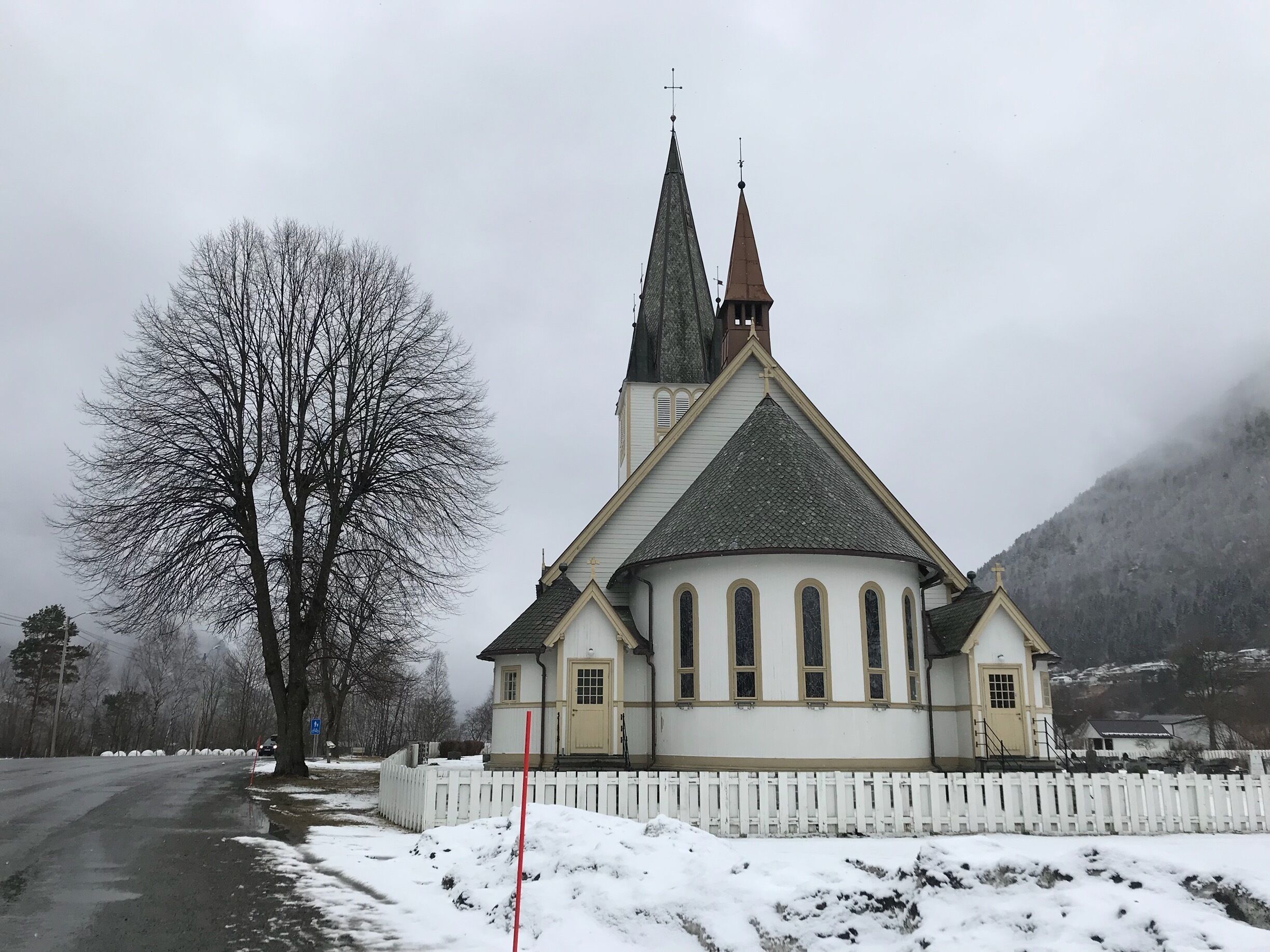 A church with a steep roof and two spires, surrounded by snow-covered ground and a bare tree.