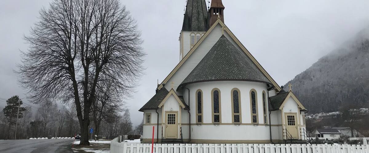 A church with a steep roof and two spires, surrounded by snow-covered ground and a bare tree.