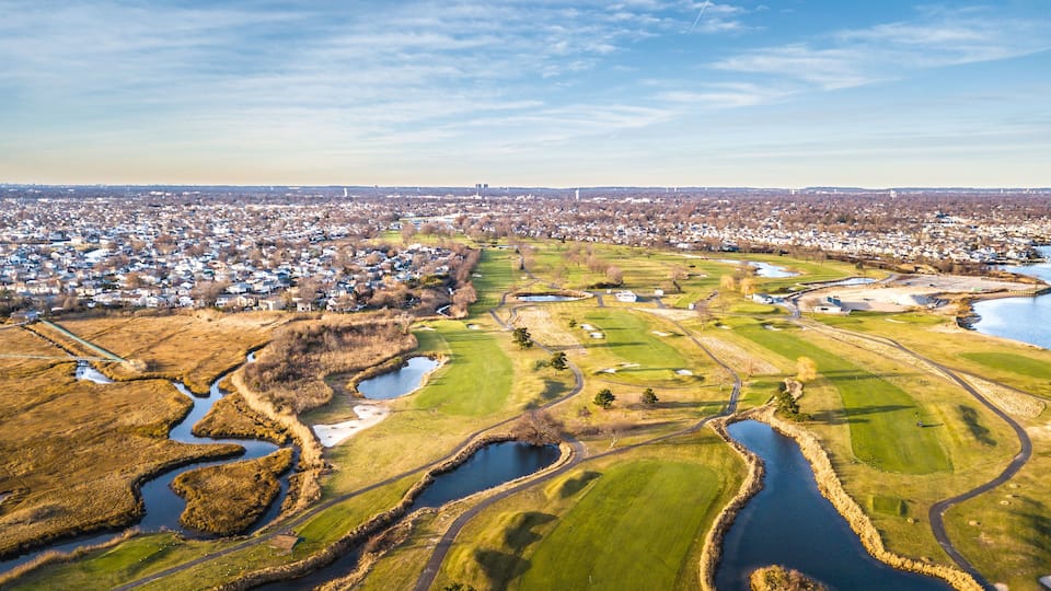 A golf course with water hazards, surrounded by a residential area and a distant city skyline.