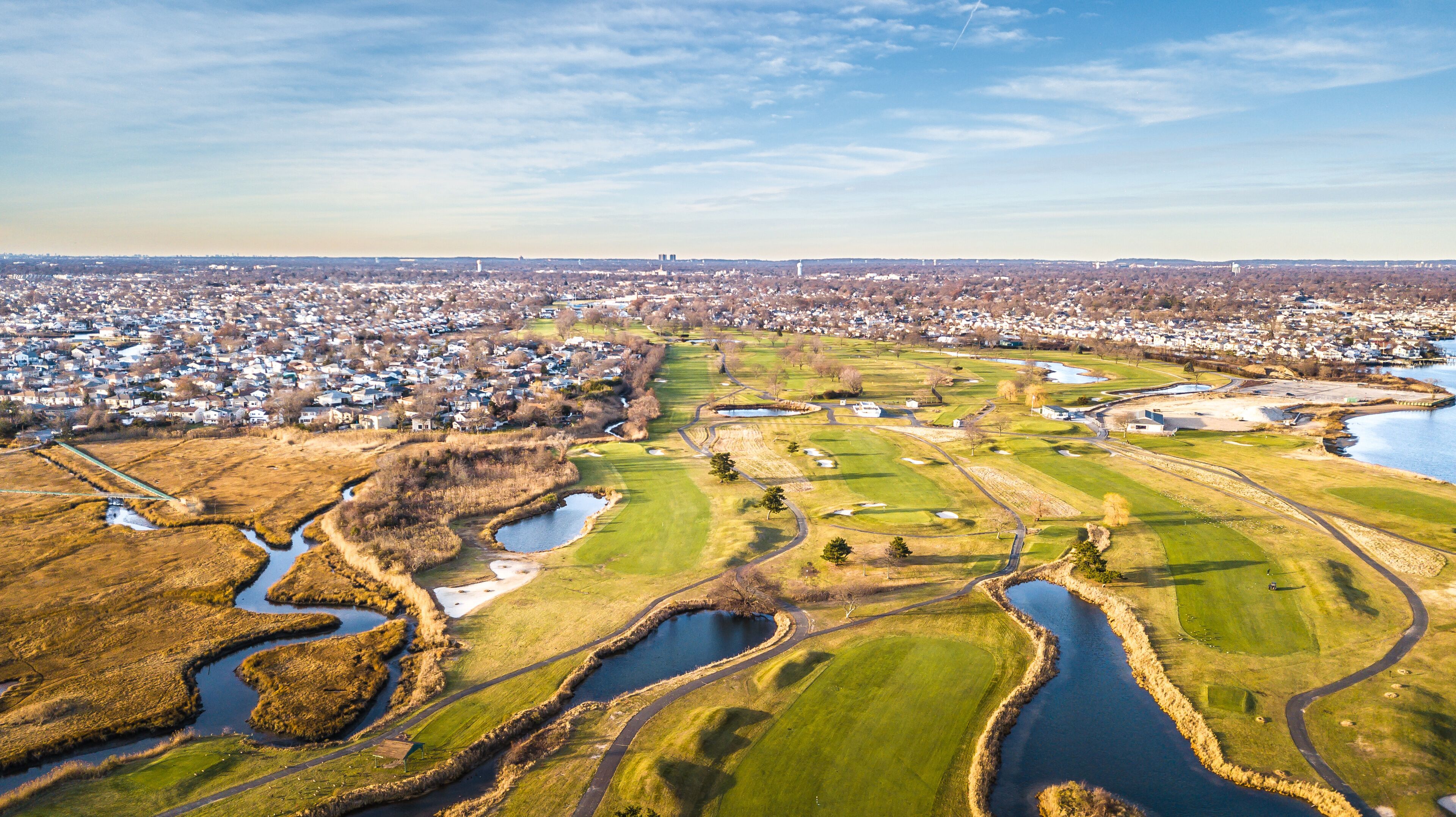 A golf course with water hazards, surrounded by a residential area and a distant city skyline.