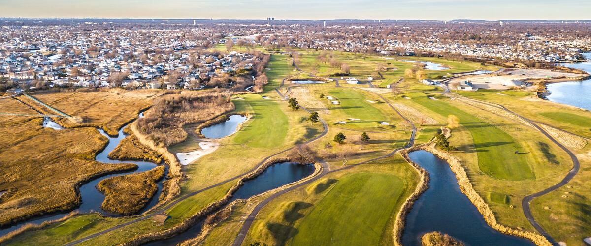 A golf course with water hazards, surrounded by a residential area and a distant city skyline.