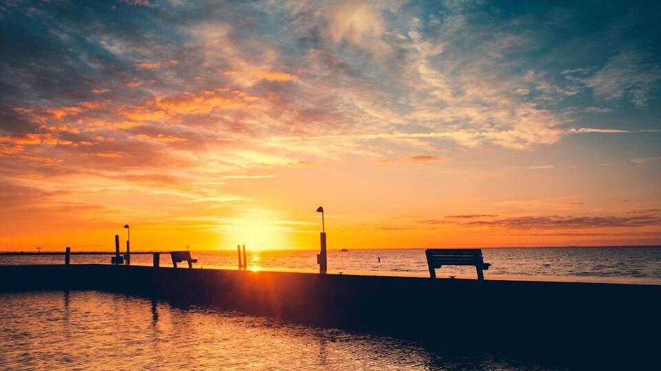 A sunset over a calm sea with a pier and benches.